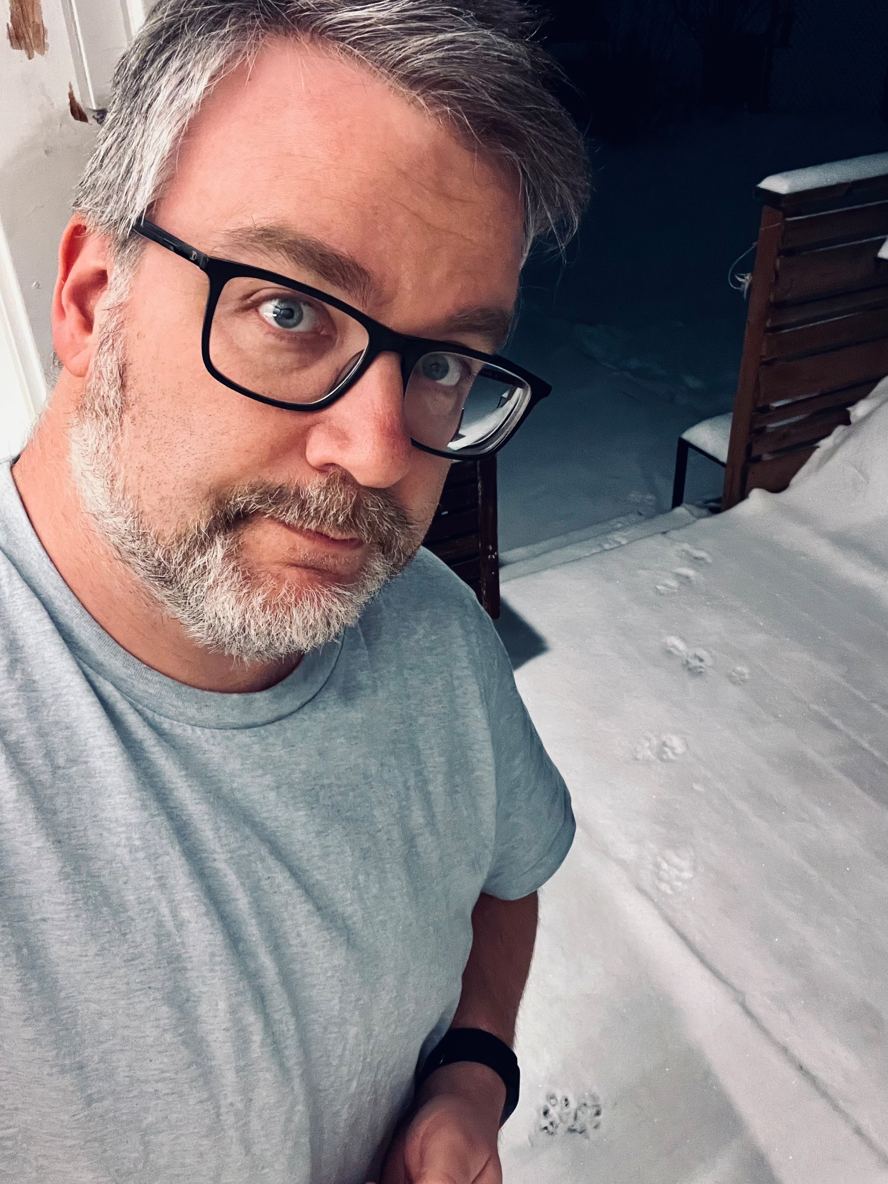 A person with glasses and facial hair is standing outside on a snowy evening next to animal footprints.