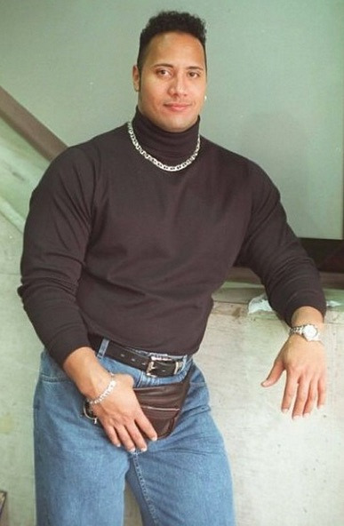 The Rock stands confidently in front of a staircase, wearing a black turtleneck, jeans, a silver chain, and a fanny pack.