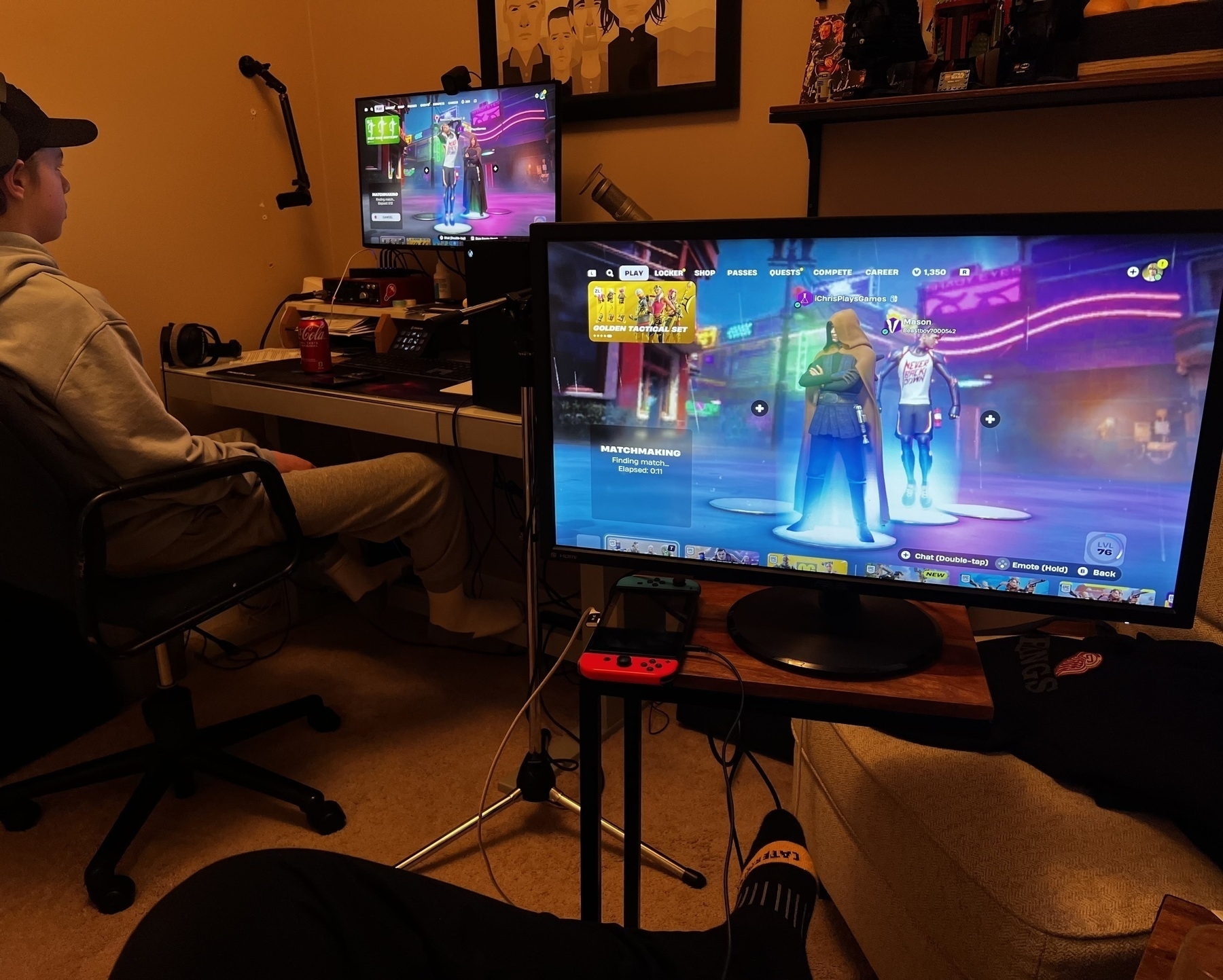 A teen sits at a computer monitor with an Xbox in the background, while a switch connected to a monitor is in the foreground. 