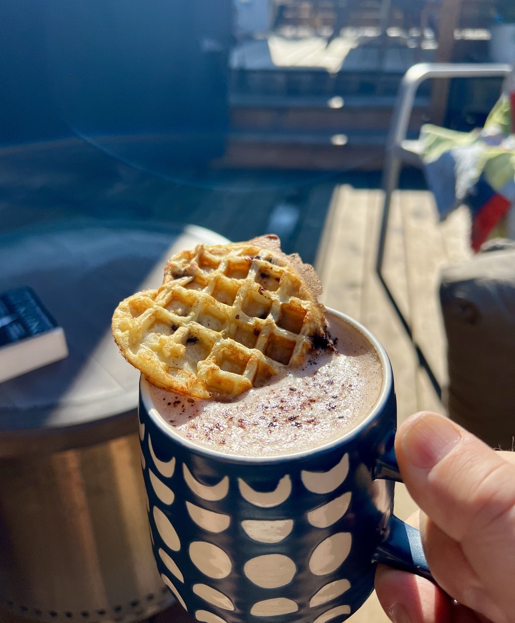 A mug of coffee with a small heart shaped waffle sitting half in the coffee is being held on a deck. 