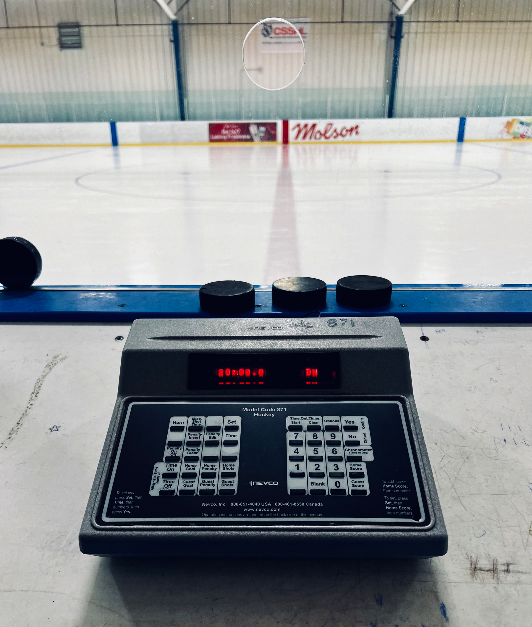 A digital scoreboard and control panel face an empty ice rink with advertisement banners in the background.