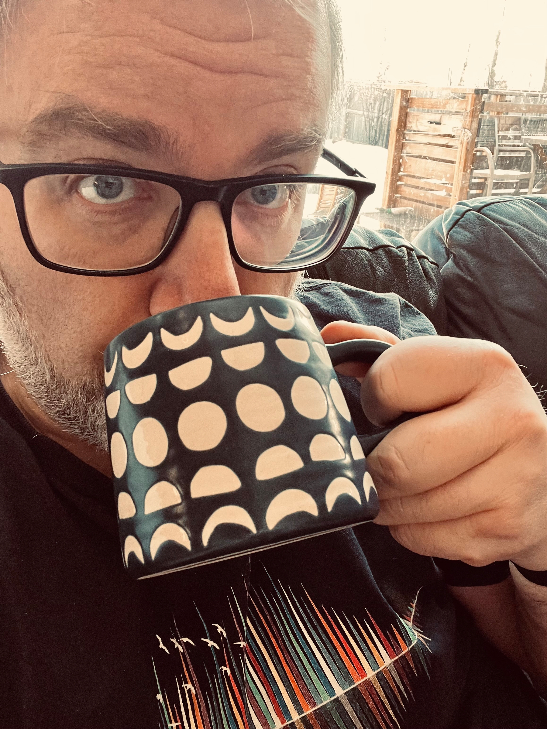 A person wearing glasses is sipping from a mug with a black and white geometric pattern.